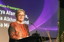 Foto: Eine Frau mit kurzen, grauen Haaren steht an einem Rednerpult. Im Hintergrund ist ein Bildschirm mit Text und violettem Grafikdesign.