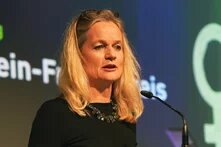 Foto: Eine Frau mit langen, blonden Haaren spricht in ein Mikrofon. Im Hintergrund ist ein Bildschirm mit teilweise lesbarem Text.